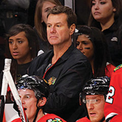 Mike Gapski looks forward as he stands behind sitting Chicago Blackhawks players