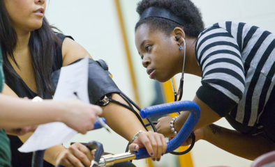 African American female student with stethescope check blood pressure of classmate on stationary bike